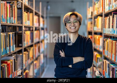 Ritratto di sorridente studente asiatico in piedi con le braccia incrociate in biblioteca del college Foto Stock