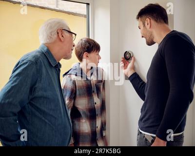 Figlio, padre e nonno che controllano il termostato intelligente sulla parete in cucina Foto Stock