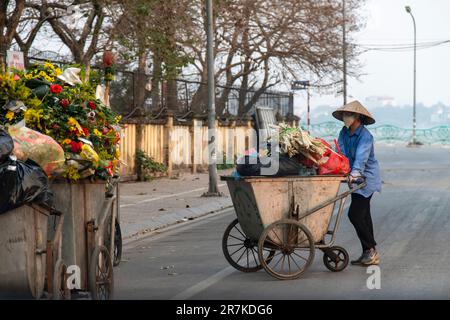 Hanoi, Vietnam-aprile 2023; primo piano di una donna vietnamita che spinge un carrello spazzatura in strada sormontato da rifiuti tra cui fiori Foto Stock