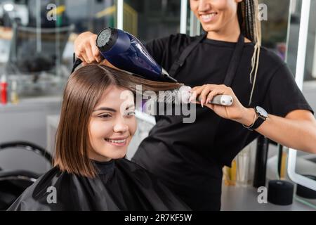 positività, salone blow dry, professionale parrucchiere con spazzola rotonda e asciugacapelli styling capelli di una cliente femminile, felice bruna donna con corto Foto Stock