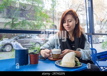 giovane donna latina di etnia colombiana, seduta a colazione nella caffetteria prendendo la tazza di caffè dal tavolo, con il cibo sul tavolo Foto Stock