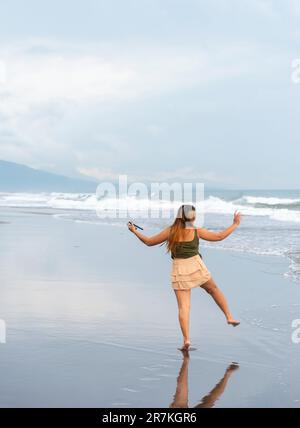 Tenendo il suo telefono per selfie, su una spiaggia vicino Manila al tramonto, aimlessly passeggiando le calde acque costiere, lungo le sabbie lisce, avendo divertimento, ballando aroun Foto Stock