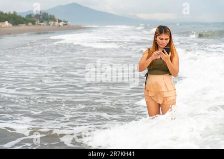 Tenendo il suo telefono per selfie, su una spiaggia vicino Manila al tramonto, aimlessly passeggiando le calde acque costiere, lungo le sabbie lisce, avendo divertimento, ballando aroun Foto Stock