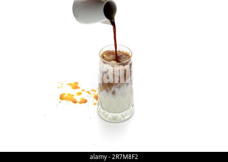 Il caffè viene versato in un bicchiere di latte ghiacciato su una superficie bianca con macchie di caffè Foto Stock