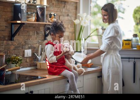 Giovane ragazza che gode di un delizioso muffin in una mano mentre tiene il suo amato peloso orso giocattolo nell'altra. Al suo fianco si trova la madre premurosa, dilige Foto Stock