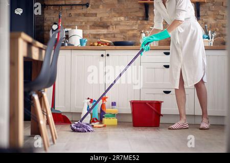 Foto senza volto di giovane donna che indossa guanti, pulizia in cucina, moping il pavimento, con bottiglie chimiche sullo sfondo. Casa, pulizia, lifes Foto Stock