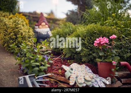 Primo piano ripresa orizzontale di giardino fiorito con guanti e attrezzi da giardinaggio, fiori, vaso di fiori, e un nano sfocato sullo sfondo. Conce di giardinaggio Foto Stock