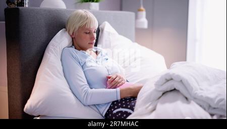 Young Woman With Hot Water Bottle On Stomach Lying In Bed Stock Photo