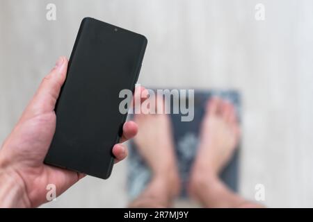Un uomo si trova sulla bilancia. Il ragazzo sta pesando se stesso. Bilance da pavimento per la salute. Controllo del peso. Schermo verde dello smartphone. Foto Stock