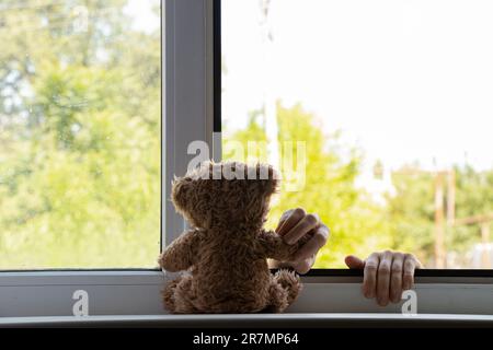 Le mani dei bambini si tengono sulla finestra dal lato della strada e tengono un orsacchiotto, suicidio, mani fuori dalla finestra, suicidio Foto Stock