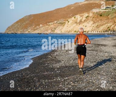 Un uomo maturo degli anni sessanta corre lungo la costa di una spiaggia soleggiata, godendosi il suono delle onde che si avvolgono Foto Stock