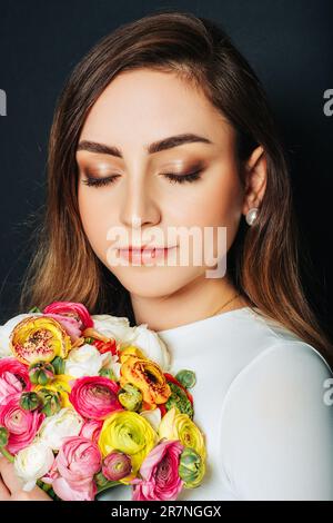 Studio primo piano ritratto di bella giovane donna con capelli lunghi e luminosi, trucco professionale, vestito bianco, tenendo colorati fiori buttercup Foto Stock