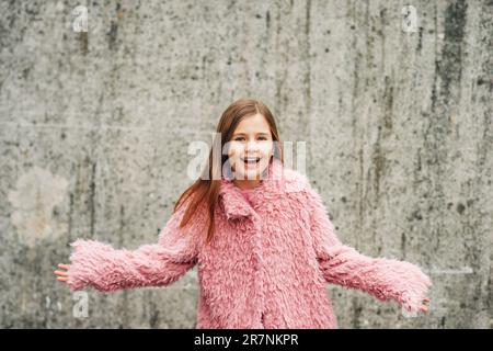 Ritratto all'aperto di una bambina eccitata che indossa un cappotto rosa di pelliccia sintetica, che si posa contro le mura grigie della città, braccia aperte Foto Stock