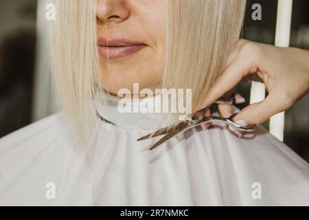 Il primo piano di un parrucchiere taglia i capelli bianchi bagnati di un cliente in un salone. Il parrucchiere taglia una donna. Vista laterale di una mano che taglia i capelli con le forbici Foto Stock