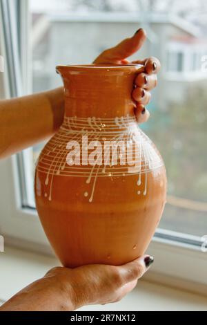 caraffa in terracotta smaltata bianca in mani femminili. primi piatti in ceramica su sfondo sfocato. verticale. Foto Stock