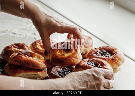 la mano femminile prende un panino da un piatto pieno di cheesecake appena sfornati con formaggio cottage sul tavolo della cucina. cottura in forno. preparazione della cottura Foto Stock