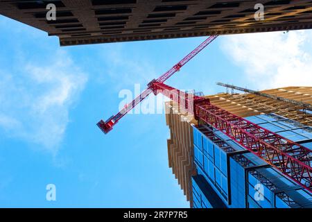 Vista maestosa di una gru rossa alta che opera contro un cielo blu brillante. Ombre e riflessi e una prospettiva impressionante aggiungono un tocco di interesse visivo. Foto Stock