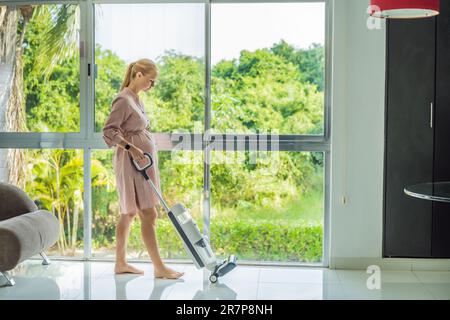 La giovane donna incinta ama pulire la sua casa. Facile pulizia con un aspirapolvere wireless. Donna incinta che pulisce il pavimento con l'aspirapolvere portatile Foto Stock