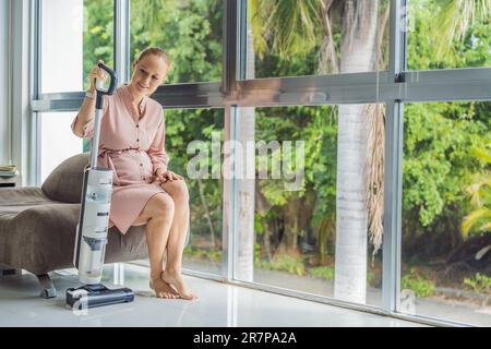 La giovane donna incinta ama pulire la sua casa. Facile pulizia con un aspirapolvere wireless. Donna incinta che pulisce il pavimento con l'aspirapolvere portatile Foto Stock