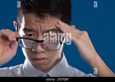 L'uomo asiatico tiene i suoi templi, sperimentando un intenso mal di testa. Un momento difficile per lui. Raffigurato su sfondo blu Foto Stock