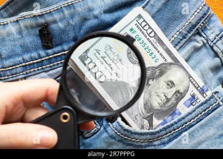 Cento dollari americani che escono da un buco in jeans primo piano ingrandito con una lente d'ingrandimento per la lettura Foto Stock