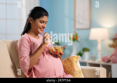 Donna incinta indiana felice che mangia Macedonia di frutta mentre si siede sul divano a casa - concetto di gravidanza nutrizione, alimentazione sana e benessere Prenatale. Foto Stock