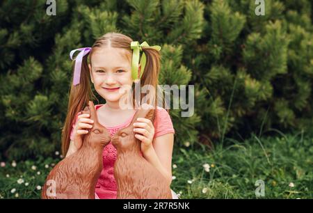 Ritratto all'aperto di una simpatica bambina che gioca in un giardino con due coniglietti di cioccolato Foto Stock