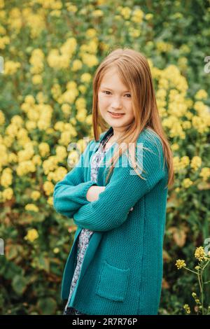 Ritratto primaverile all'aperto di bella ragazza di 6-7 anni che gioca in campo di fiori gialli, indossando cardigan verde, braccia incrociate Foto Stock