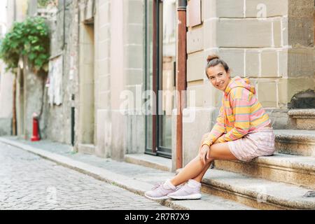 Ritratto all'aperto di giovane donna graziosa seduta sui gradini della città, indossando felpa a strisce rosa e gialla, gonna, comode sneaker viola Foto Stock