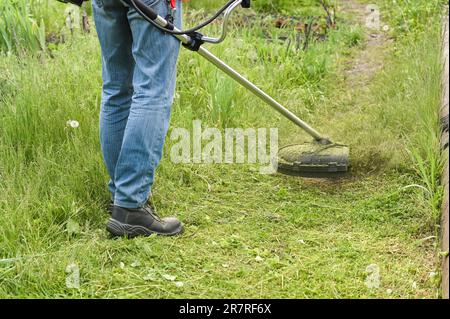 Lavoratore che taglia erba alta con tagliaerba elettrico o a benzina nel parco cittadino o nel cortile. Processo di rifilatura del prato con rasaerba manuale Foto Stock