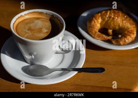 Primo piano di una tazza di ceramica ripiena di caffè appena fatto, accompagnata da alcuni deliziosi croissant, su un piattino bianco Foto Stock