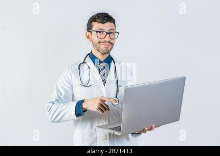 Medico sorridente che usa e punta computer portatile isolato. Giovane medico in occhiali utilizzando il computer portatile e guardando la macchina fotografica. Bel medico che usa un computer portatile isolato Foto Stock
