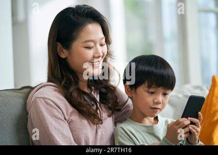 giovane madre asiatica seduta sul divano di famiglia mantenendo la compagnia con il figlio di cinque anni Foto Stock