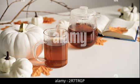 Primo piano di teiera di vetro con tè caldo, tazza, libro, foglie di acero arancione, zucche e ramo su tavolo bianco. Il concetto di inizio autunno, caduta atm Foto Stock