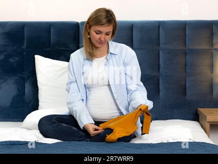 Happy Pregnent Woman ha uno starter kit arancione, un set di tuta unisex a manica lunga, seduto su un letto blu. La donna indossa abiti casual, bianco e blu Foto Stock