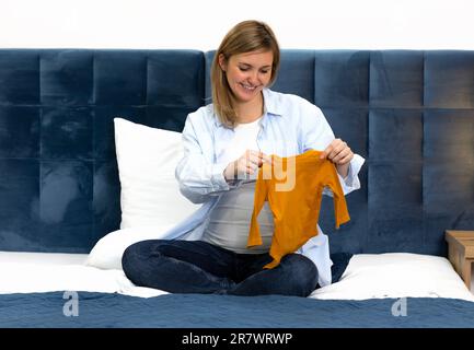 Happy Pregnent Woman ha uno starter kit arancione, un set di tuta unisex a manica lunga, seduto su un letto blu. La donna indossa abiti casual, bianco e blu Foto Stock