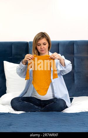 Happy Pregnent Woman ha uno starter kit arancione, un set di tuta unisex a manica lunga, seduto su un letto blu. La donna indossa abiti casual, bianco e blu Foto Stock