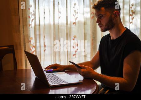 Uomo ispanico che studia utilizzando un multischermo con computer portatile e smartphone vicino alla finestra Foto Stock