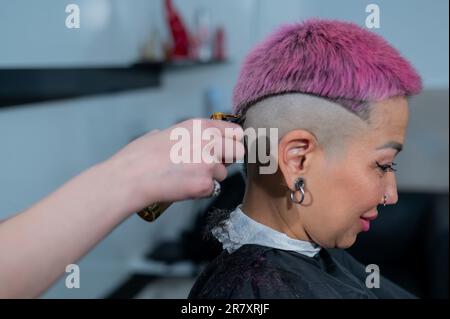 Il parrucchiere rasa il tempio di una cliente femminile. Donna asiatica con capelli corti rosa in barbiere. Foto Stock