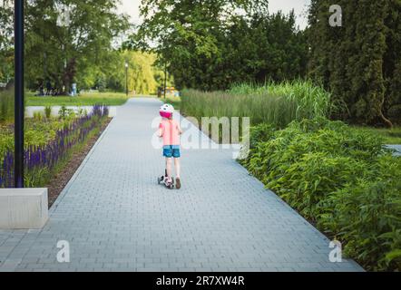 Una bambina guida uno scooter in un bellissimo parco pieno di verde. Foto Stock