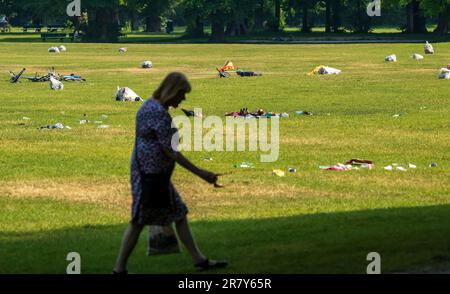 Monaco, Germania. 18th giugno, 2023. Una donna passa davanti al grande prato nel Giardino Inglese, dove i visitatori del giorno e della notte precedenti hanno lasciato dietro grandi quantità di rifiuti. Decine di bottiglie di birra e vino, vetri rotti, sacchetti di snack e altri rifiuti sono sdraiati intorno allo spazio aperto. Sullo sfondo, i sacchetti di spazzatura già cotti sono pronti per la rimozione. Credit: Peter Kneffel/dpa/Alamy Live News Foto Stock