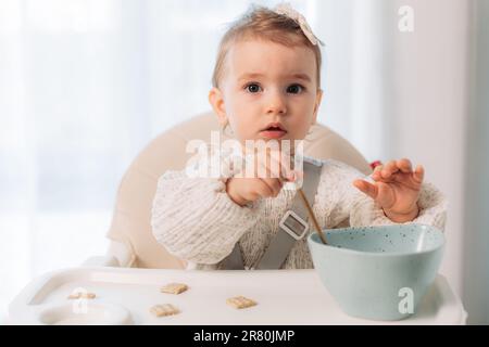 Bambina seduta in un seggiolone mangiando da sola con un cucchiaio Foto Stock