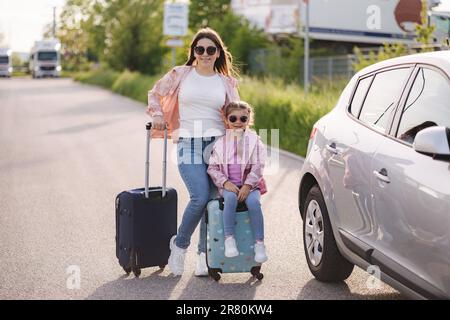 Adorabile bambina si siede sulla valigia del suo bambino e aspetta il viaggio con sua mamma. Due belle ragazze preparano Foto Stock