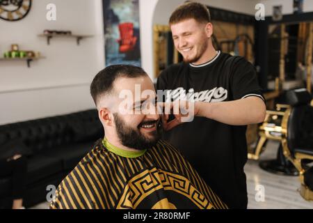 Uomo fiducioso barbuto in visita hairstylist nel barbiere. Lavoro berbero professionale Foto Stock