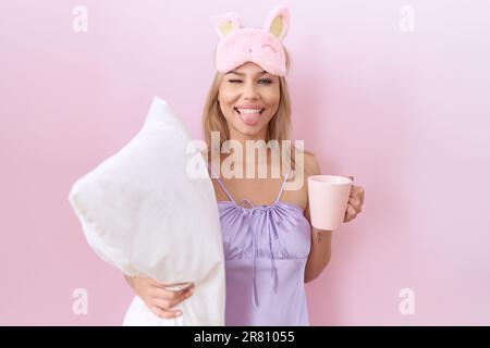 Giovane donna caucasica che indossa pajama che abbraccia il cuscino e bere caffè che si attacca lingua fuori felice con l'espressione divertente. Foto Stock