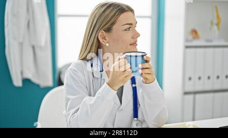 Giovane donna bionda medico sorridente sicuro bere caffè in clinica Foto Stock