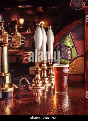 La pinta di bitter servita al bar in legno superficie di una calda e tipica e tradizionale inglese invitante public house, con birra lager e maniglie della pompa dietro Foto Stock