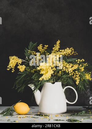still life - mimosa gialla, caraffa bianca e limone, su sfondo grigio Foto Stock