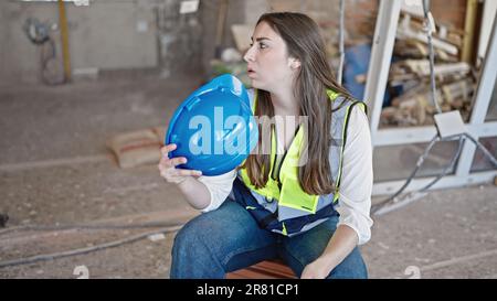 Giovane bella donna ispanica costruttore stanco utilizzando l'hardHat come un ventilatore a mano sul sito di costruzione Foto Stock