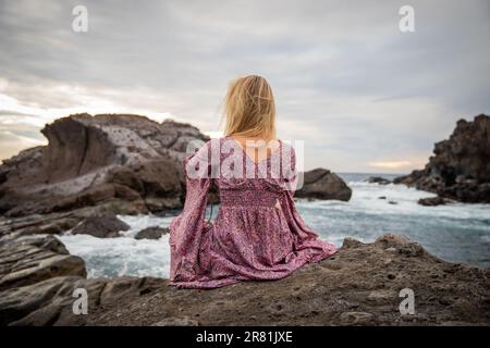 Una donna seduta sulle rocce guarda l'oceano con le onde Foto Stock